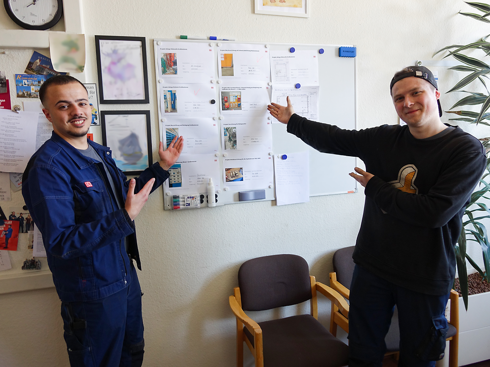 Zwei junge Männer zeigen auf ein Poster an der Wand
