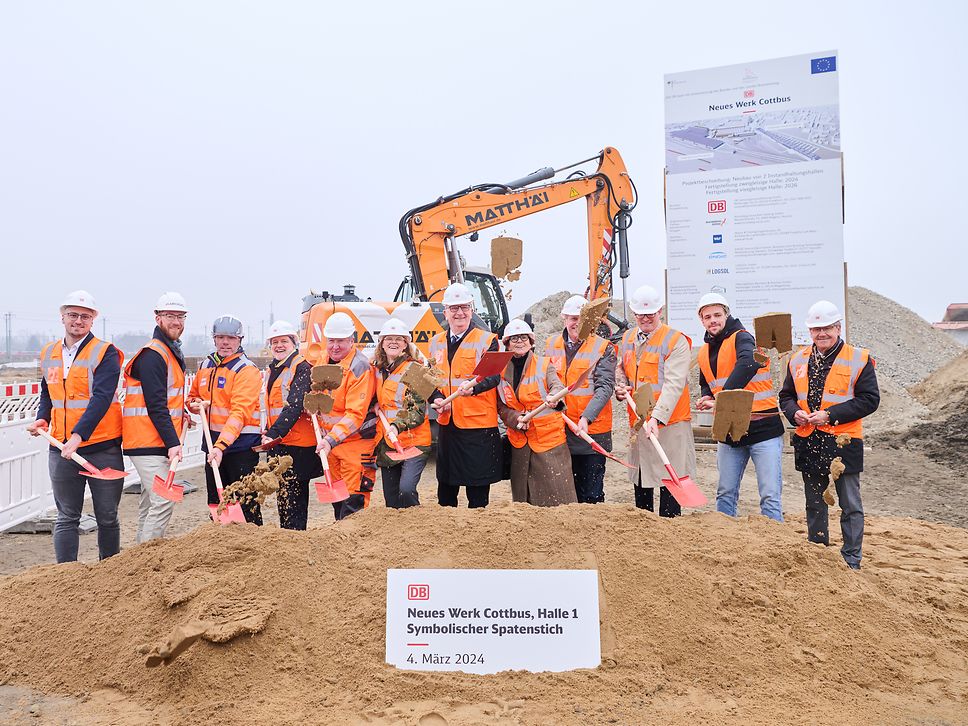 12 people stand in front of a sandheap performing the symbolic ground-breaking ceremony for Hall 1 of the new Cottbus factory.