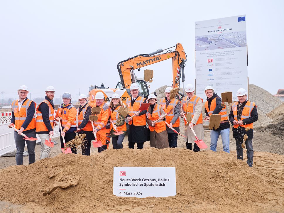 12 Personen stehen vor einem Sandhügel und führen den symbolischen Spatenstich für die Halle 1 des neuen Werk Cottbusses durch.